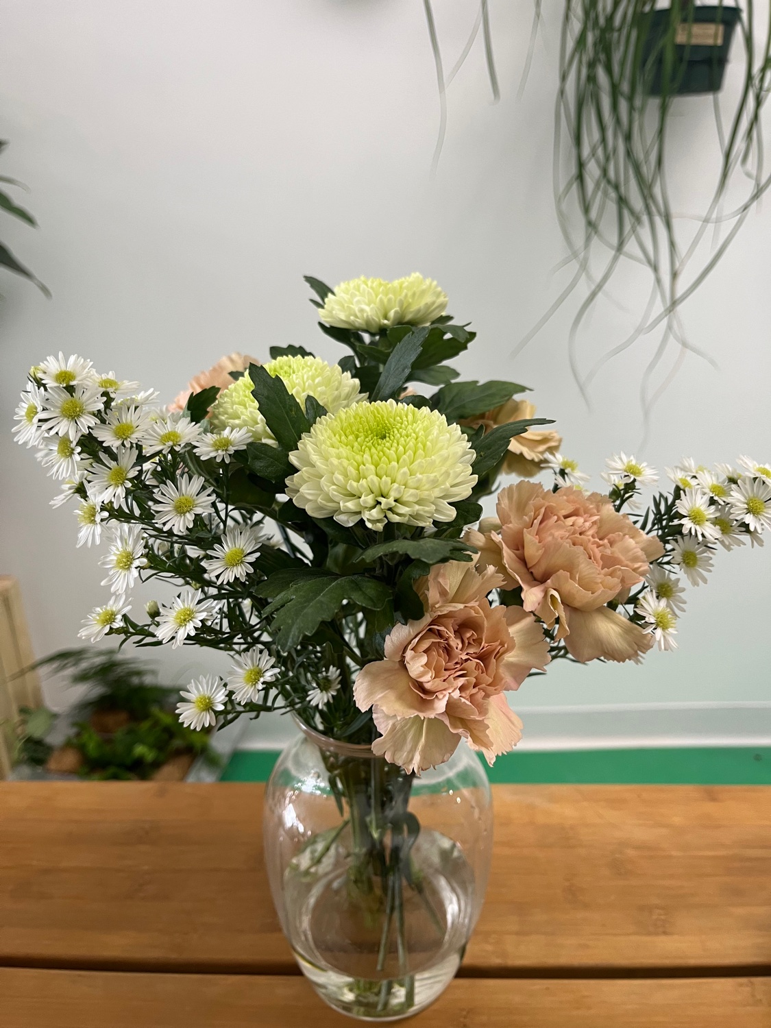 Small weekly bouquet in a clear glass vase with pale green mums, peach carnations, and white daisies