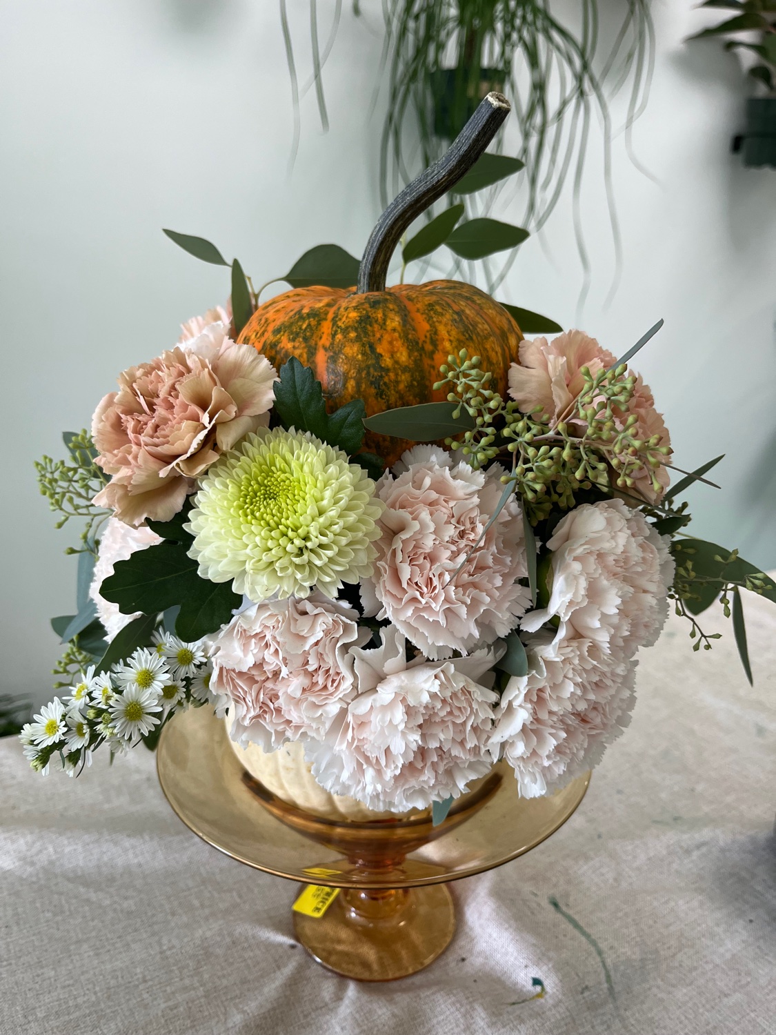 Seasonal pumpkin arrangement with blush carnations, a pale green mum, and a small pumpkin, displayed on an amber glass stand